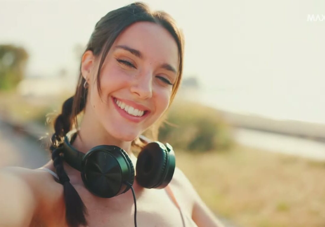 A smiling woman with headphones round her neck takes a selfie on a sunny path